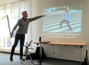 Filmmaker Rick Minnich points to a screen where an image of him filming on a staircase is showing. Below is the text: BUNT Stiftung Video Workshop